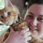 1000020573jpg ⋆ Sassy Koonz Maine Coon Cattery Close-up of a smiling person holding an orange tabby kitten with a colorful collar and a bell visible on its neck