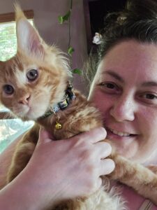 1000020573jpg ⋆ Sassy Koonz Maine Coon Cattery Close-up of a smiling person holding an orange tabby kitten with a colorful collar and a bell visible on its neck