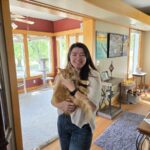 IMG_6736jpeg ⋆ Sassy Koonz Maine Coon Cattery Smiling woman in a white sweater holding an orange cat inside a sunlit living room with wooden trim.