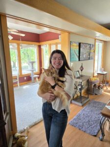 IMG_6736jpeg ⋆ Sassy Koonz Maine Coon Cattery Smiling woman in a white sweater holding an orange cat inside a sunlit living room with wooden trim.